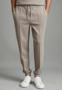 Pantaloni de trening bej cu talie cu șnur, buzunare laterale și detalii cu cusături verticale, asociați cu adidași albi pe un fundal neutru.