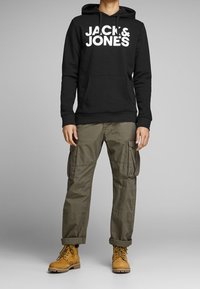 Schwarzer Kapuzenpullover mit großem weißen "JACK & JONES" Logo, kombiniert mit olivgrünen Cargo-Hosen und braunen Arbeitsstiefeln.