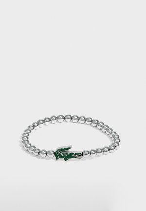 Bracelet en perles argentées avec un pendentif logo crocodile vert au centre sur fond blanc.