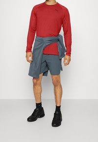 Rotes langärmliges Sportshirt, graue Shorts, grauer Pullover um die Taille gebunden und schwarze Turnschuhe. Der Stoff scheint glatt, leicht und figurbetont zu sein.