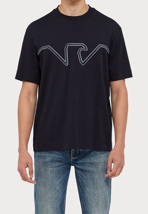 Homme portant un T-shirt noir à manches courtes avec un dessin d'aigle en contour blanc sur la poitrine et un jean bleu.