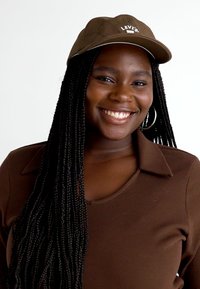 Brun baseballcap med "LEVI'S"-logo, båret med en brun krageoverdel. Flettet hår henger ned og skaper en teksturert kontrast.
