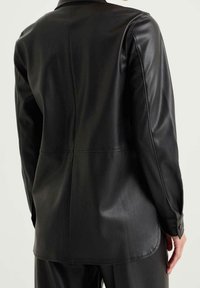 Bakåtsikt av en person som bär en långärmad svart läderjacka med knäppningsmanchetter och matchande svarta byxor.