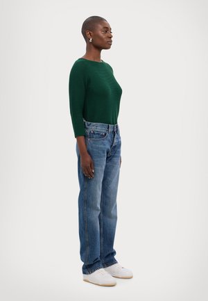 Grønn ribbet langermet topp, høy midje blå denim jeans, hvite joggesko. Modell som står sideveis, og viser frem den avslappede passformen og design detaljer.