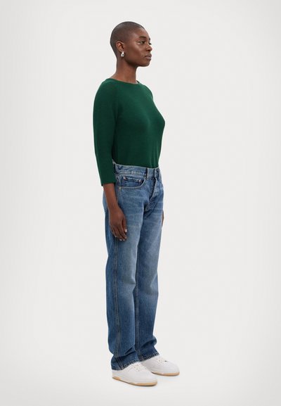 Haut vert à rayures, à manches longues, jean bleu en denim taille haute, baskets blanches. Modèle de profil, mettant en avant la coupe décontractée et les détails de conception.