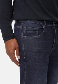Jeans en denim bleu foncé avec une coupe slim, dotés d'une poche avant, de surpiqûres contrastées et d'une fermeture éclair. Associés à une chemise noire à manches longues.