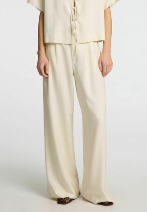 Completo beige chiaro a due pezzi composto da una maglietta a maniche corte con chiusura a fiocco e pantaloni a gamba larga. Tessuto morbido e liscio con un design minimal.