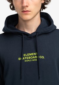 Navy hoodie met gele logotekst, voorzien van trekkoorden en een zachte textuur. Focus op casual streetwear ontwerp.