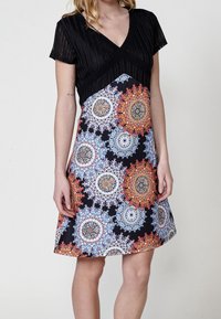 Femme portant une robe noire à motifs mandalas circulaires colorés sur la jupe, sur un fond uni.