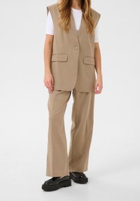 Beige maßgeschneiderte Weste ohne Ärmel, mit einem Knopfverschluss und zwei vorderen Taschen, kombiniert mit passenden weit geschnittenen Hosen und schwarzen Plateauschuhen.