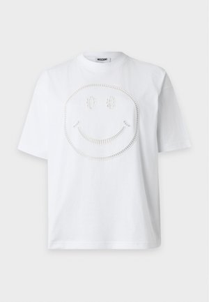 Femme portant un t-shirt blanc avec un visage souriant, une jupe midi beige à boutons, des talons noirs, tenant un sac à main rouge sur un fond clair uni.