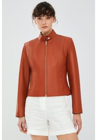Bruine leren jas met een hoge kraag en een gouden rits, voorzien van gestructureerde naden en twee zijzakken. Draagt met witte shorts.