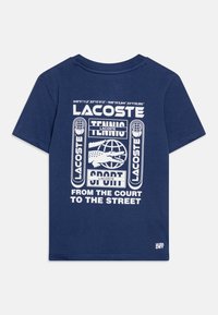 Navyblauwe katoenen t-shirt met een witte grafische ontwerpelementen, inclusief de tekst "LACOSTE," een wereldbol en tennisgerelateerde afbeeldingen. Etiket "ULTRA DRY" op de zoom.