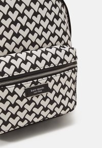 Svart och vit ryggsäck med hjärtformade mönster. Har en dragkedjeficka och en etikett med "kate spade." Strukturerad form och texturerad tyg.