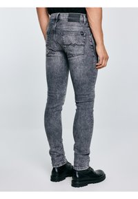 Mees, kes kannab kitsaid kulunud musta teksapükse ja musti nahast saapaid, seisab ühe käega vabalt külje kõrval tavalise heleda tausta ees.