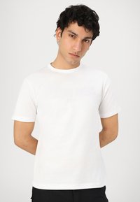 T-shirt bianco in cotone con maniche corte, scollatura rotonda e sottile design in rilievo del testo. Texture liscia, vestibilità casual.
