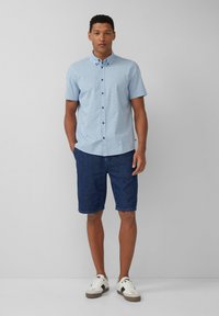 Chemise légère bleue à manches courtes avec un motif géométrique, assortie à un short en denim foncé et des baskets blanches avec des accents noirs.