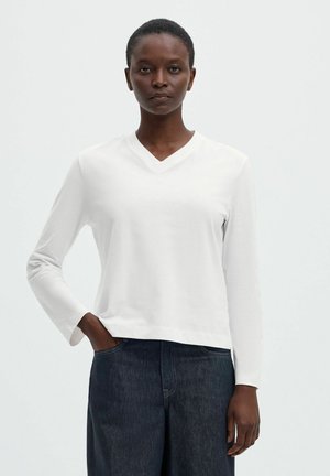 Personne portant un haut blanc uni à manches longues avec col en V et un pantalon en jean bleu foncé, debout avec une main dans la poche devant un fond clair.
