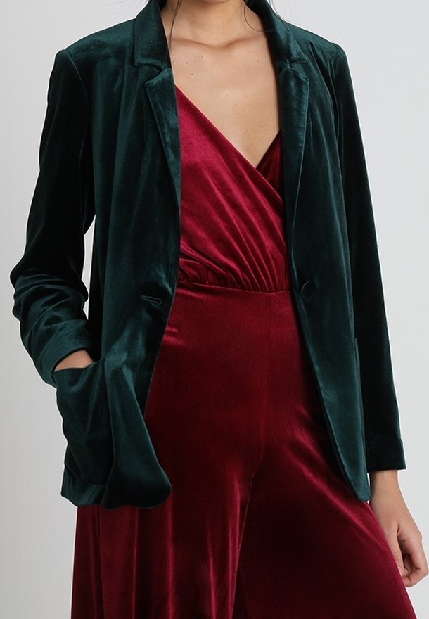 Femme portant un blazer en velours vert foncé sur une combinaison cache-cœur en velours rouge profond, une main dans la poche et fond neutre.