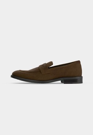 Mocassin homme en daim marron avec petit talon noir et détail de bride sur le dessus, vu de profil sur fond uni.