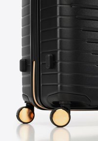 Esquina de maleta rígida negra con bordes horizontales y cuatro ruedas giratorias con detalles dorados sobre fondo blanco.