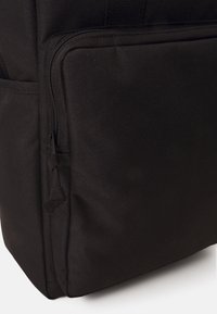 Sac à dos noir avec une surface texturée, présentant une grande poche zippée sur le côté et des coutures robustes. Le matériau semble durable et pratique.