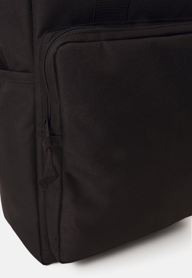 Schwarzer Rucksack mit strukturierter Oberfläche, der eine markante seitliche Reißverschlusstasche und robuste Nähte aufweist. Das Material wirkt langlebig und praktisch.