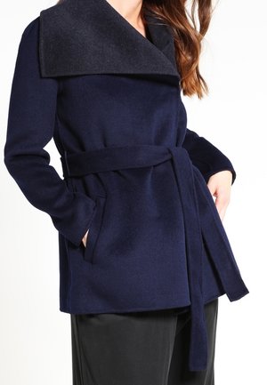 Femme portant un manteau bleu marine ceinturé avec un grand col noir, les mains dans les poches, assorti à un pantalon noir.