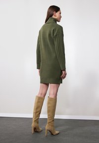 Robe tricotée vert olive avec un col haut et des manches côtelées, accompagnée de bottes beiges à talons hauts jusqu'aux genoux. Tissu texturé, design ajusté.