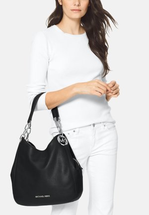 Mujer vestida completamente de blanco sosteniendo un gran bolso negro Michael Kors con asas de cadena plateada y colgante con el logo.