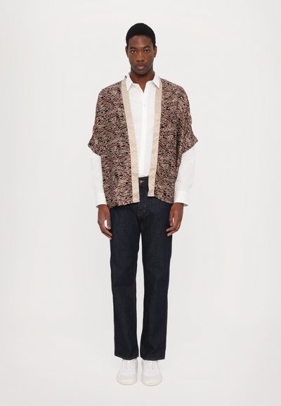 Veste de style kimono à motifs bruns avec des accessoires beiges, portée sur une chemise blanche ; associée à un jean en denim foncé et des baskets blanches.
