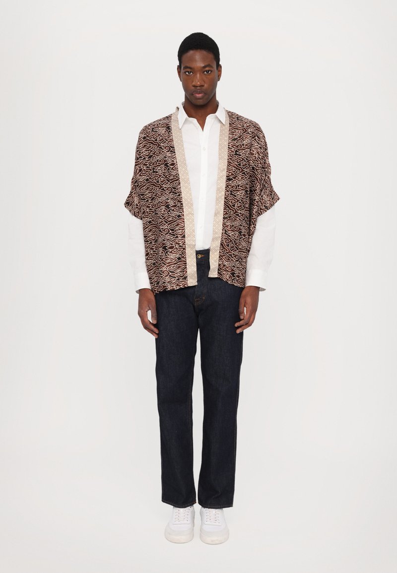 Brunmønstret kimono-stil jakke med beige kant, båret over en hvit skjorte; kombinert med mørk denimjeans og hvite joggesko.