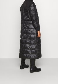 Lång svart pufferjacka med vertikal quiltad design, glänsande nylonmaterial, ribbade manschetter och huva, matchad med kraftiga svarta stövlar.