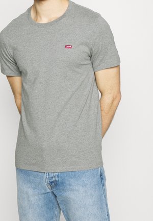 Hombre con una camiseta gris lisa de Levi's con un pequeño logo rojo en el pecho y vaqueros azul claro, con los brazos detrás de la espalda.