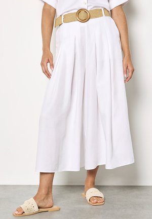 Frau trägt weiße, weit geschnittene, gekürzte Hosen mit Falten, einen beigen gewebten Gürtel mit rundem Schnallenverschluss und beigefarbene Slip-On-Sandalen.