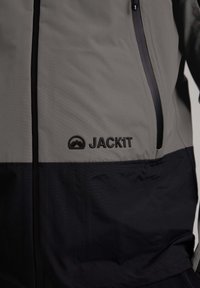 Giacca grigia e nera con zip frontale, due tasche laterali e logo "JACK1T" sul petto; tessuto liscio con un taglio moderno.