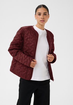 Femme portant une veste matelassée bordeaux par-dessus un t-shirt blanc et un pantalon noir, les cheveux tirés en arrière et de petites boucles d'oreilles.
