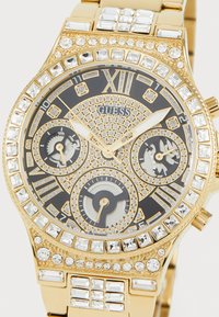 Montre dorée avec un cadran noir, des embellissements en cristal et trois sous-cadrans. Présente des chiffres romains et un bracelet en métal texturé.