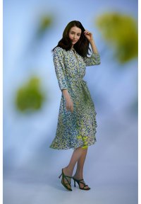 Abito midi floreale in blu, giallo e verde, con cintura in vita, maniche lunghe e orlo a volant. Abbinato a sandali verdi con cinturini.