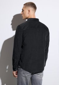 Camisa negra de corduroy con un corte relajado, mangas largas, puños con botones y un collar de punta, combinada con vaqueros gris claros.