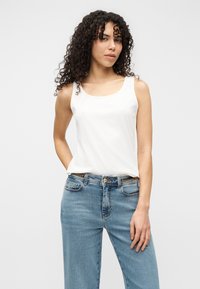 Λευκό αμάνικο τοπ με στρογγυλή λαιμόκοψη, συνδυασμένο με ανοιχτό μπλε τζιν παντελόνια φαρδιάς γραμμής με χρυσή αλυσίδα ως διακοσμητικό στη ζώνη.