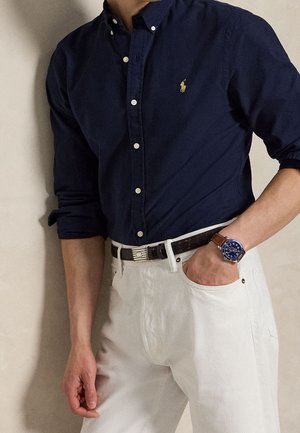 Mann trägt ein dunkelblaues Hemd mit Knöpfen, weiße Hose, braunen Gürtel und eine Armbanduhr mit blauem Zifferblatt und braunem Lederarmband.