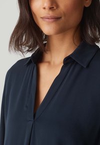 Blusa leggera blu navy con scollatura a V e colletto rilassato, realizzata in tessuto morbido e liscio, con una vestibilità leggermente drappeggiata.