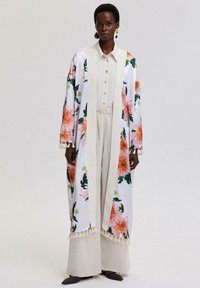 Blommig lång kimono i vitt med stora orange och gröna blommor, med en kontrasterande beige krage och fransdetaljer vid nederkanten.