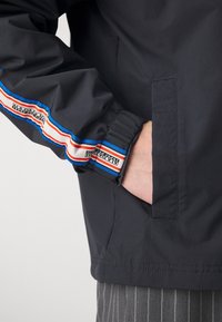 Ciemna kurtka z elastycznymi mankietami, z niebiesko-czerwonymi paskami. Posiada boczną kieszeń oraz gładką, elegancką fakturę.