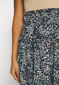 Femme portant une jupe fleurie bleu marine avec de petites fleurs roses, blanches et vertes, et une taille élastique froncée, main reposant sur le côté.