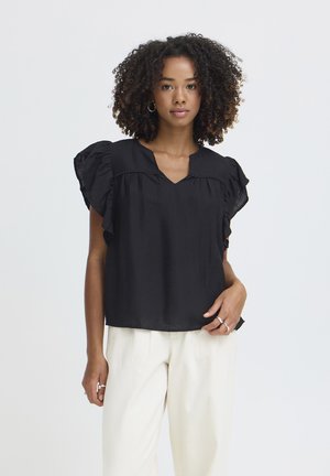 Kvinde med krøllet hår iført en sort bluse med flæser og korte ærmer samt hvide bukser, stående mod en ensfarvet lys baggrund.