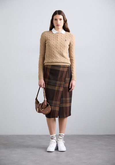 Beige cable-knit sweater with a white collared shirt, paired with a brown plaid skirt. White sneakers and a small brown handbag complete the look.
