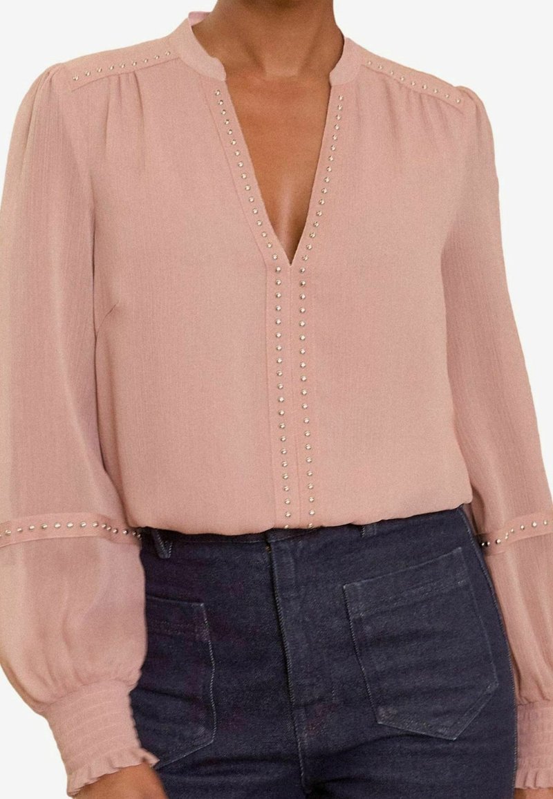 Blouse rose clair ornée de clous argentés le long de l'encolure en V, des épaules et des poignets, associée à un jean denim foncé taille haute.
