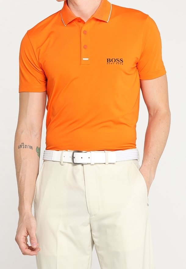 Polo orange avec col blanc et détails de boutons, arborant un petit logo noir sur la poitrine, accompagné d'un pantalon couleur crème et d'une ceinture blanche.